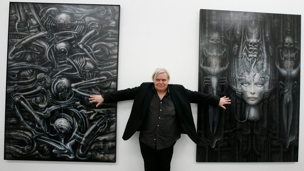 The artist H.R. Giger stands between two large paintings arms outstretched grasping the art in a black suit with a slight grin.